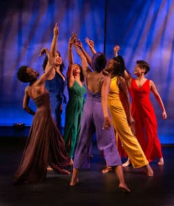 Six women dance against a purple and blue background, wearing sold colored jumpsuits